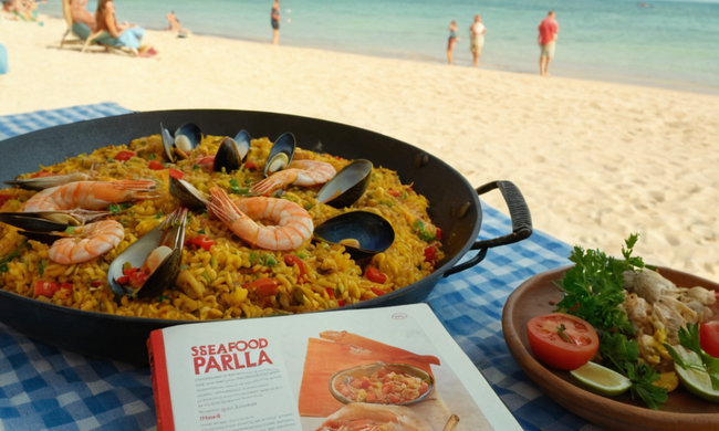 Una deliciosa paella de mariscos servida en una mesa junto a la playa, con un libro de recetas en el fondo.